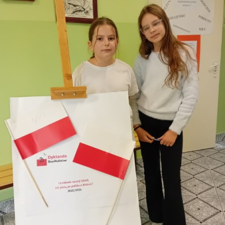 Dwie uczennice stoją przy planszy promującej Ogólnopolskie Dyktando Niepodległościowe, zdjęcie 1 z 3