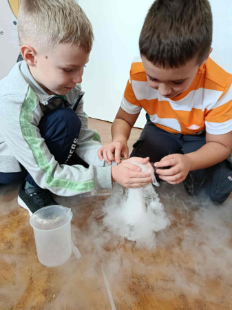 Dwóch chłopców z uśmiechem bada efekt wydobywającej się mgły nad naczyniem podczas doświadczenia chemicznego, zdjęcie 1 z 5