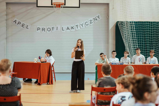 Uczennica przemawia do zgromadzonych uczniów na sali gimnastycznej, przedstawiając tematykę uzależnień, zdjęcie 2 z 5